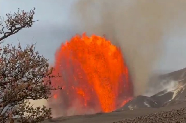 A volcanic eruption displays a large burst of bright orange lava amid smoke and ash.