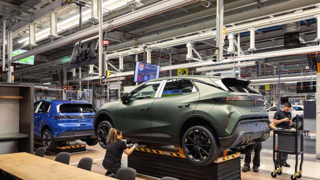 A worker inspects a green car in an automotive assembly plant with various other vehicles in the background.