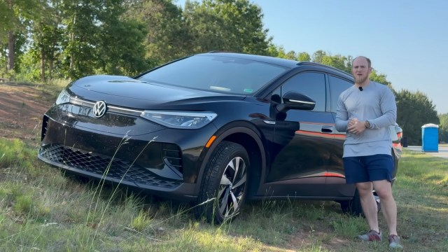 A man stands next to a black Volkswagen ID.4 in a grassy area with trees in the background.