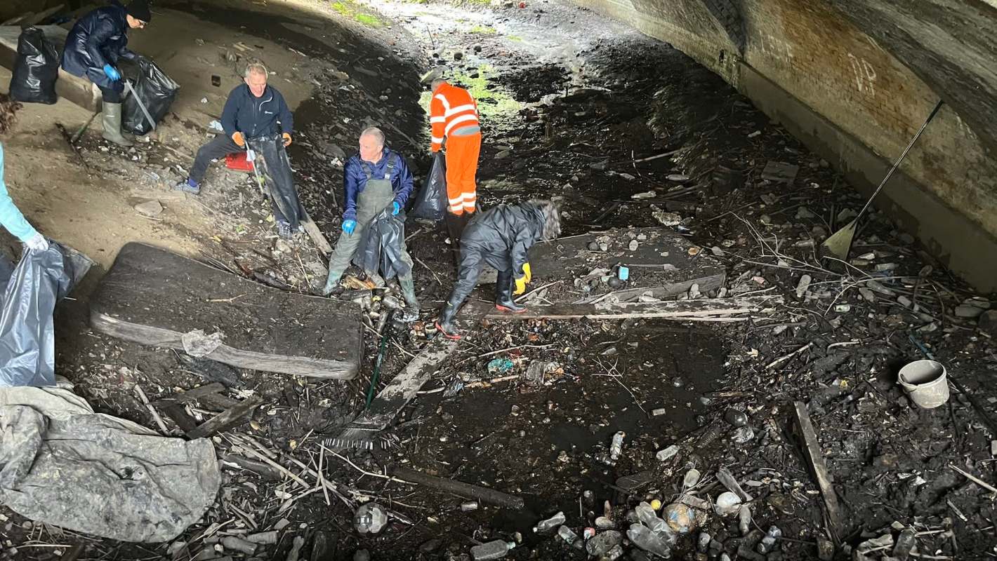 A group of volunteers collects trash in a polluted area under a bridge.