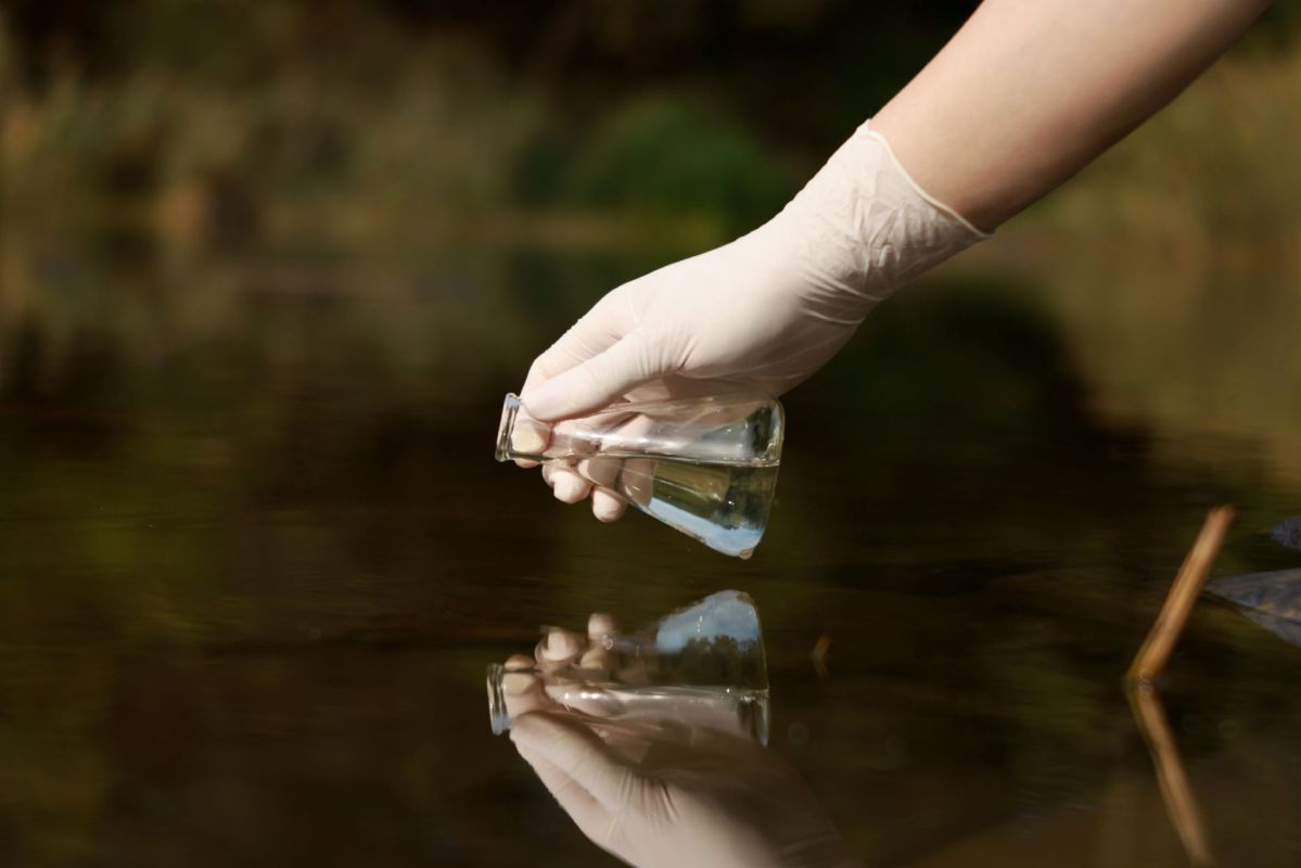 A gloved hand holds a glass vial above the water's surface.