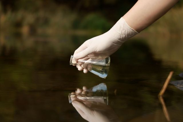 A gloved hand holds a glass vial above the water's surface.