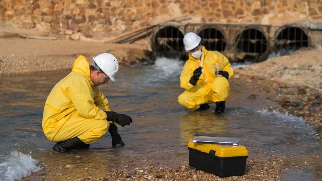 Two researchers in yellow hazmat suits collect water samples by a stream with a toolbox nearby.