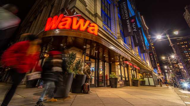 A brightly lit Wawa storefront at night with blurred pedestrians walking by.