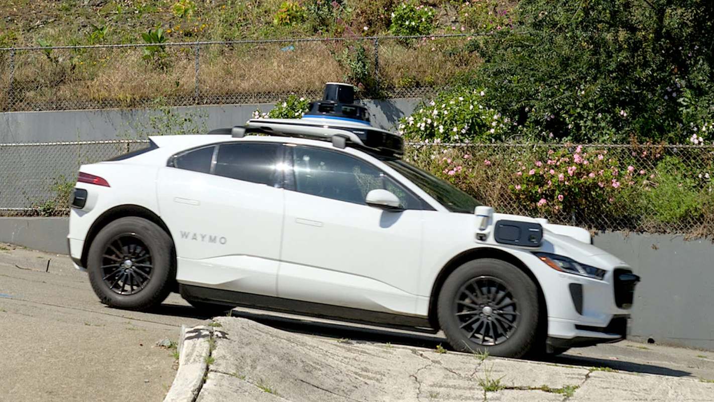 A Waymo self-driving car on a curved road next to a chain-link fence, greenery, and flowers.