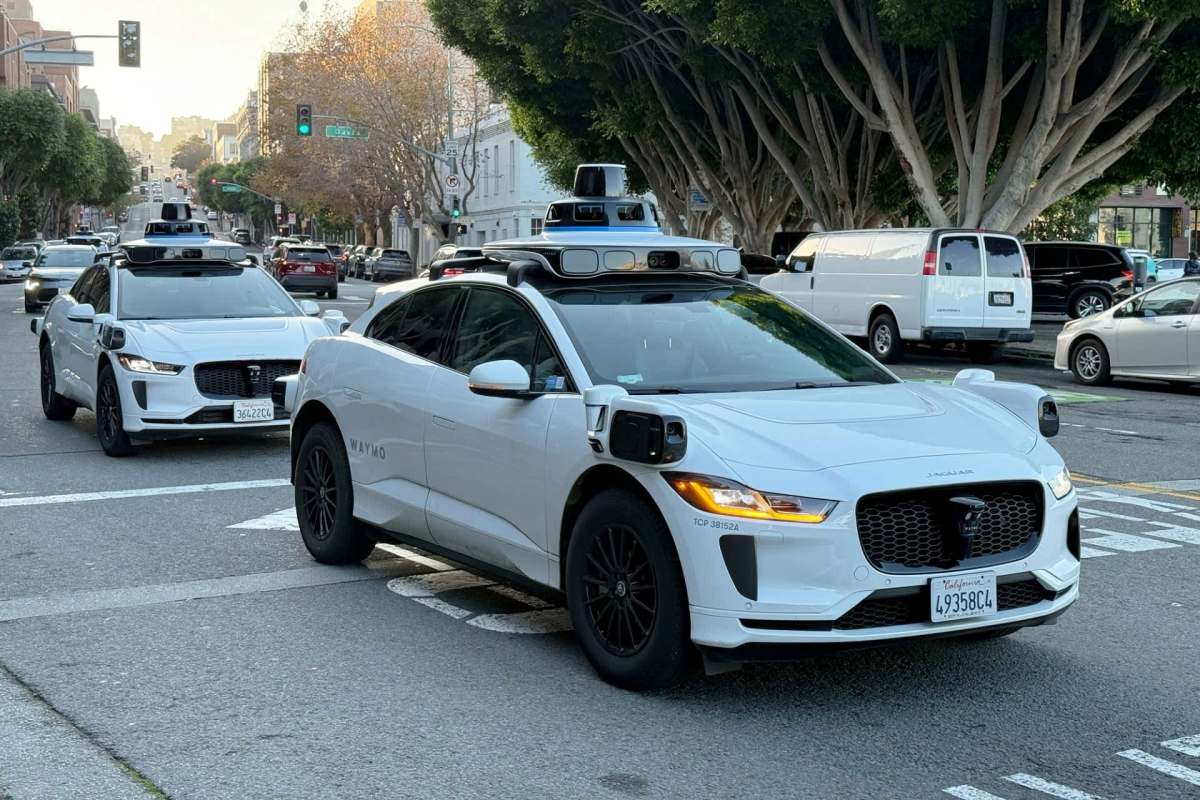 Two autonomous electric vehicles, branded with Waymo, drive on a city street lined with trees.
