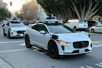 Two autonomous electric vehicles, branded with Waymo, drive on a city street lined with trees.