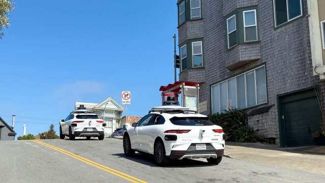 Two self-driving white cars ascend a steep street next to houses under a clear blue sky.