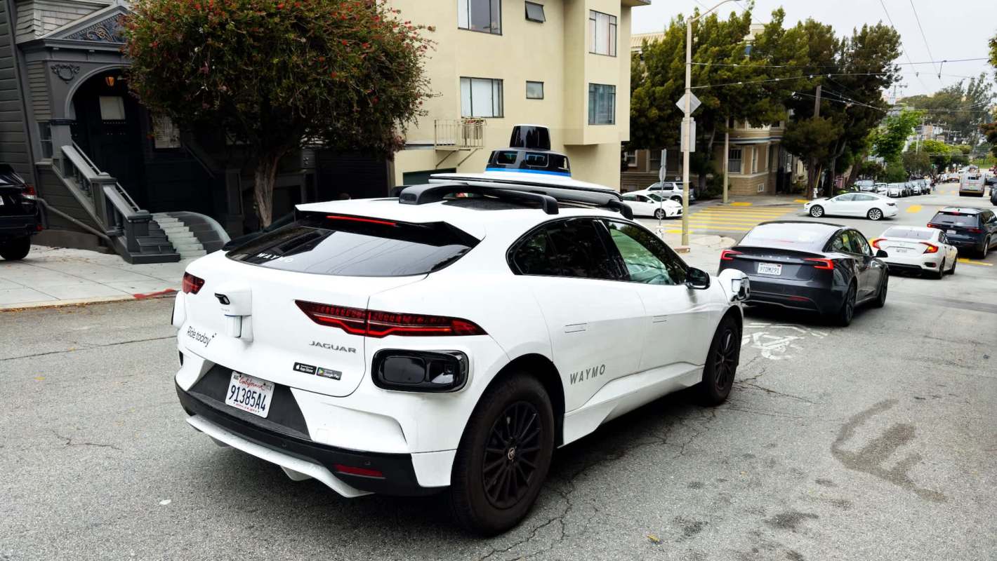 A self-driving Jaguar vehicle for Waymo with a sensor on top parked on a city street beside residential buildings.