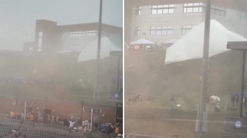 A misty scene shows a crowd by a sports venue as a large tent blows in strong winds.