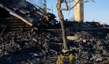 A burnt-out building with charred debris and a scorched tree against a clear blue sky.