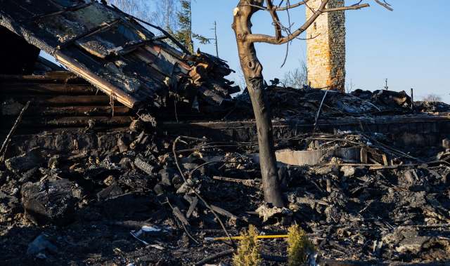 A burnt-out building with charred debris and a scorched tree against a clear blue sky.