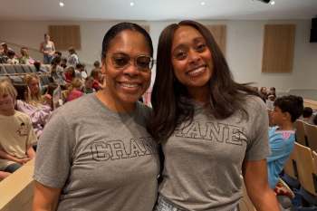 Two women wearing matching gray "CRANE" t-shirts smile at the camera in a crowded auditorium setting.