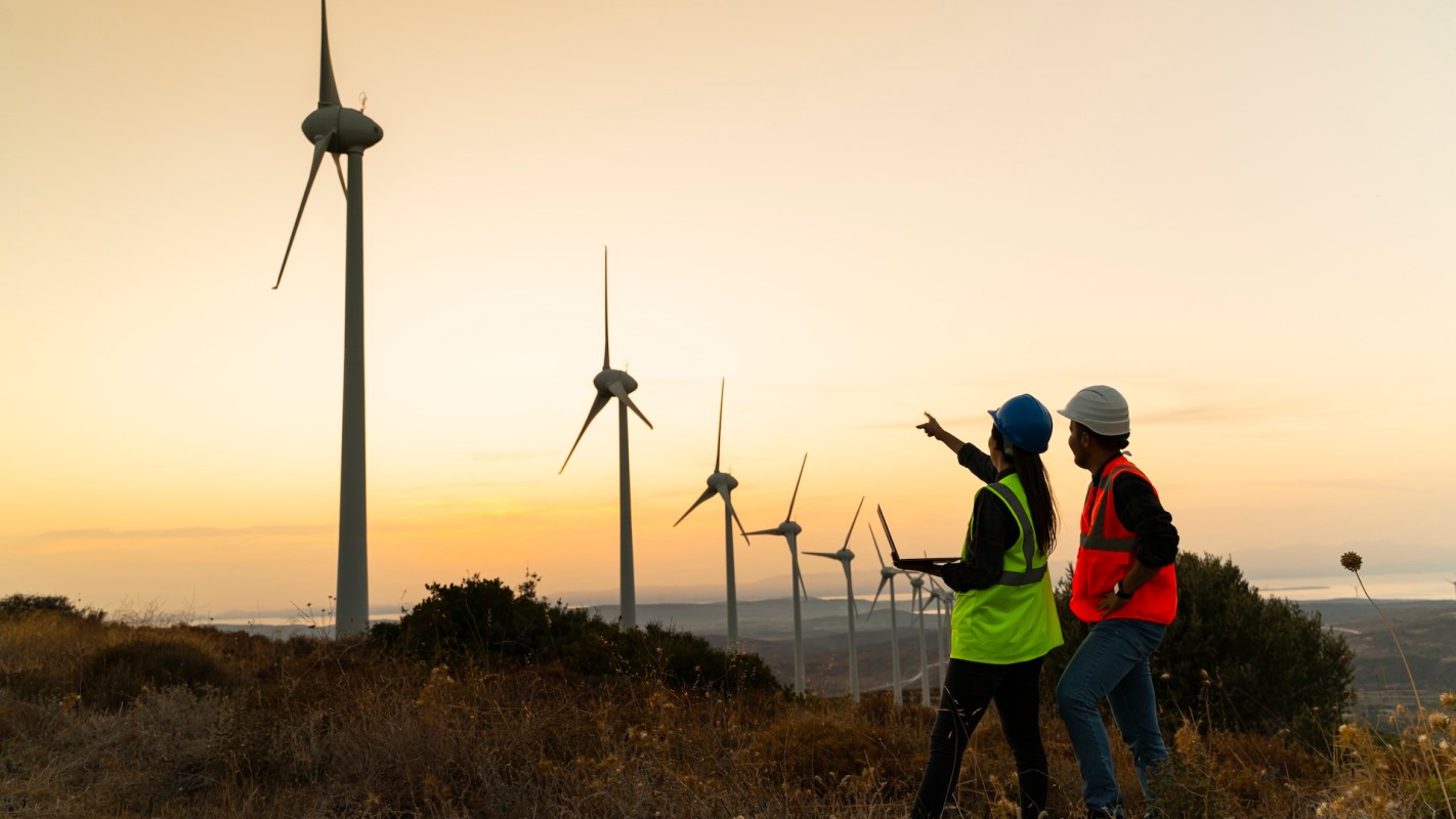 Two workers in safety gear survey wind turbines at sunset.