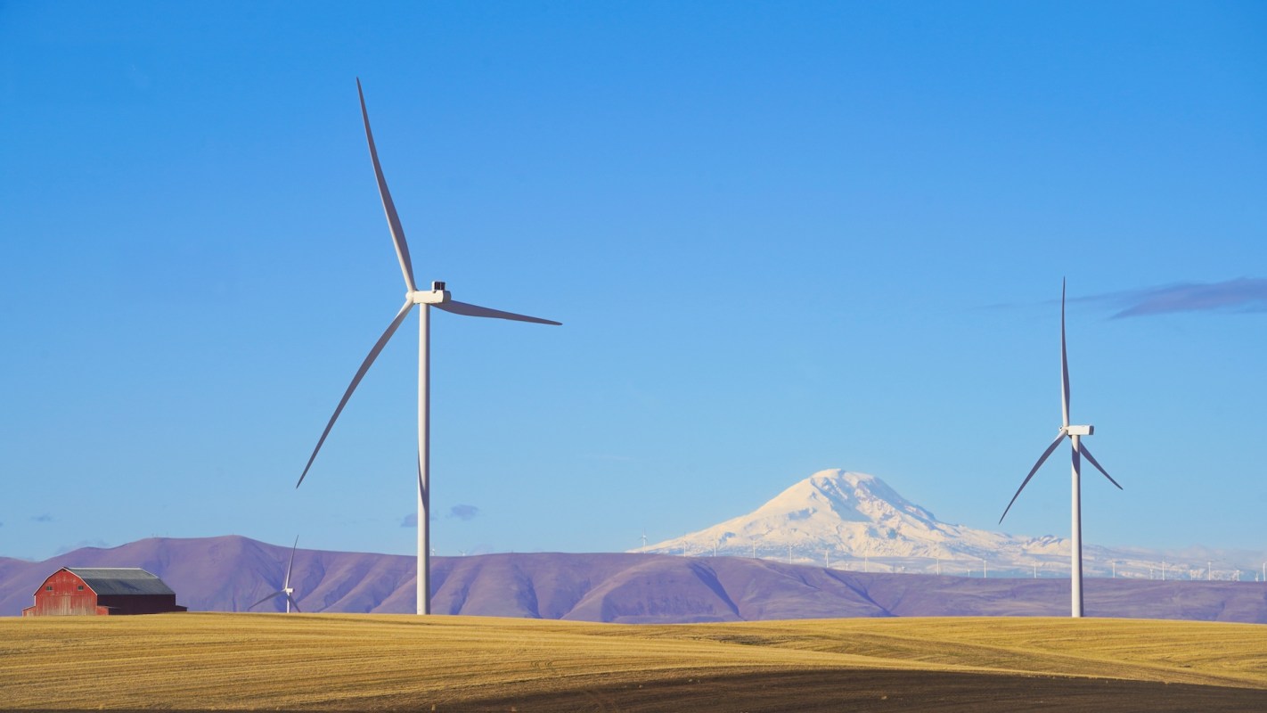 Two wind turbines stand in a golden field in front of a snow-capped mountain.