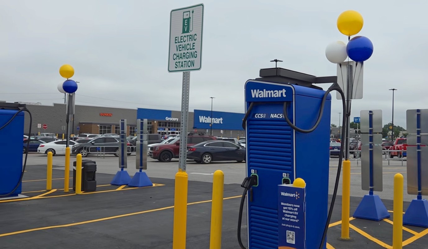 An electric vehicle charging station with a sign, located in the Walmart parking lot, surrounded by parked cars.