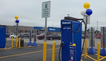 An electric vehicle charging station with a sign, located in the Walmart parking lot, surrounded by parked cars.