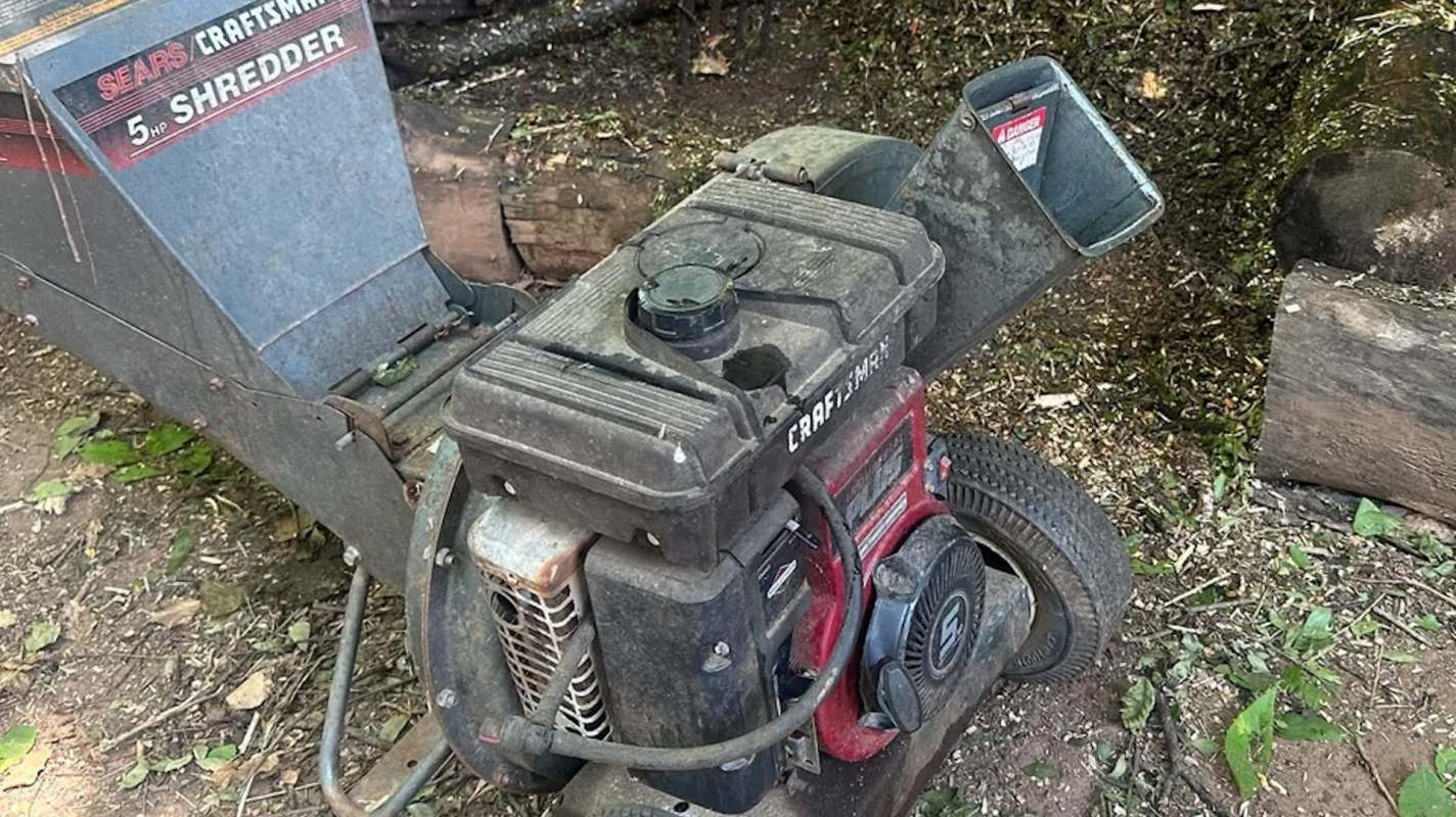 A close-up of a dusty lawn shredder machine surrounded by fallen leaves and wood debris.