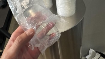 A hand holding a small clear plastic cup wrapped in crinkled film, with a metal trash can in the background.