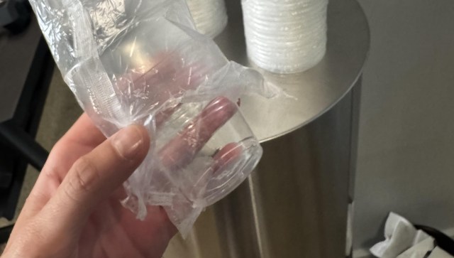 A hand holding a small clear plastic cup wrapped in crinkled film, with a metal trash can in the background.
