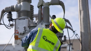 A worker in a yellow safety vest with the Ameren company logo and helmet inspects electrical equipment on a utility pole.