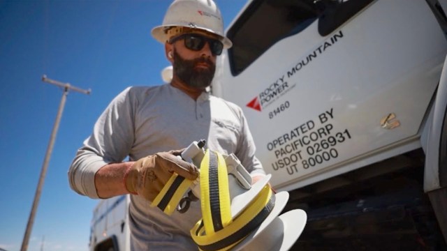 A man in a hard hat and gloves holds a spool of cable near a work truck with the Rocky Mountain Power logo.