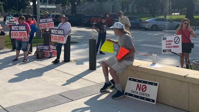 A group of protesters holding signs against data centers outside a government building on a sunny day.