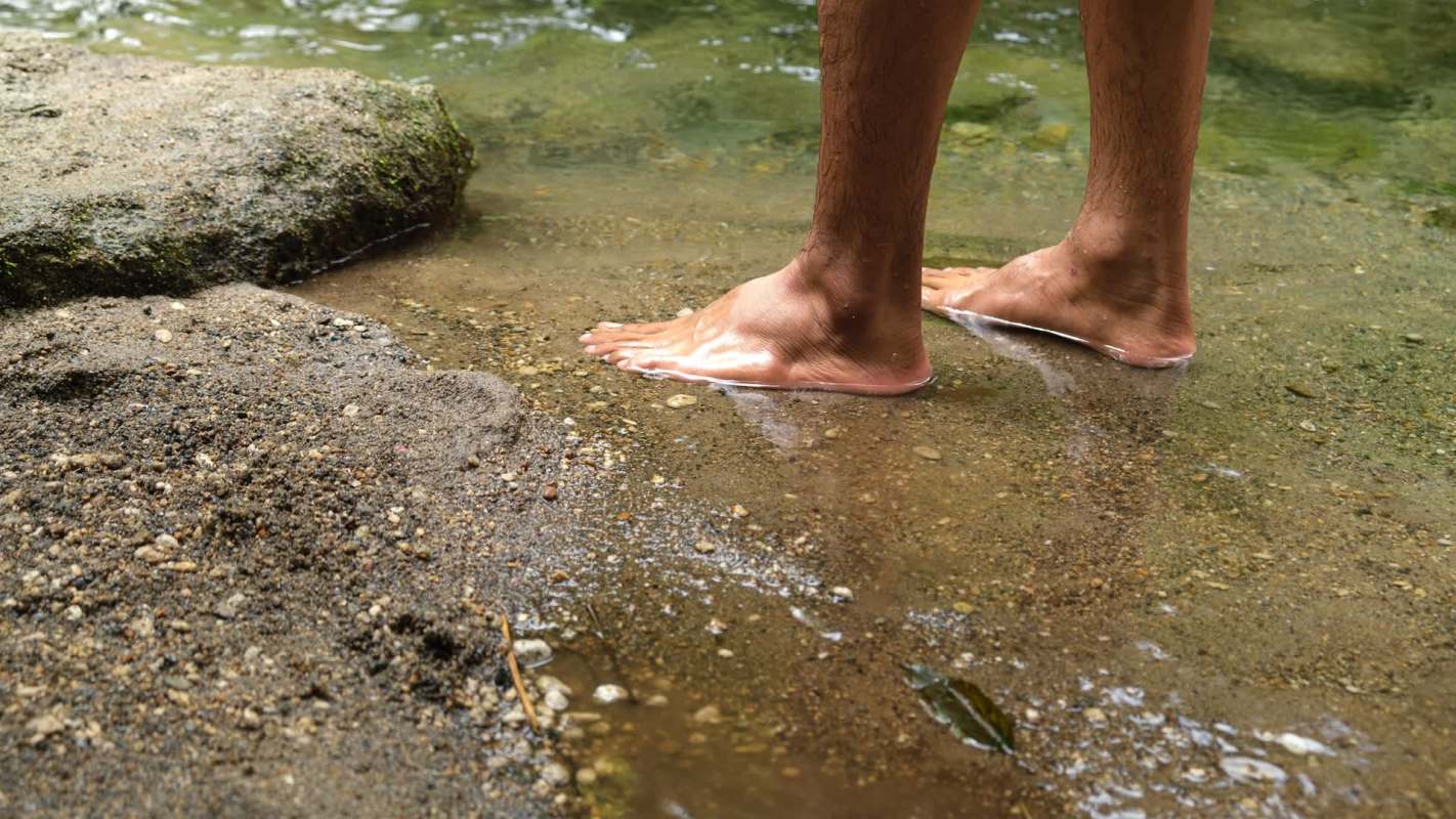 A person stands barefoot in shallow water at the edge of a river, with sand and pebbles visible around their feet.