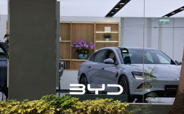 A sleek silver BYD Seal car displayed behind a glass window with the BYD logo and lush green plants in the foreground.