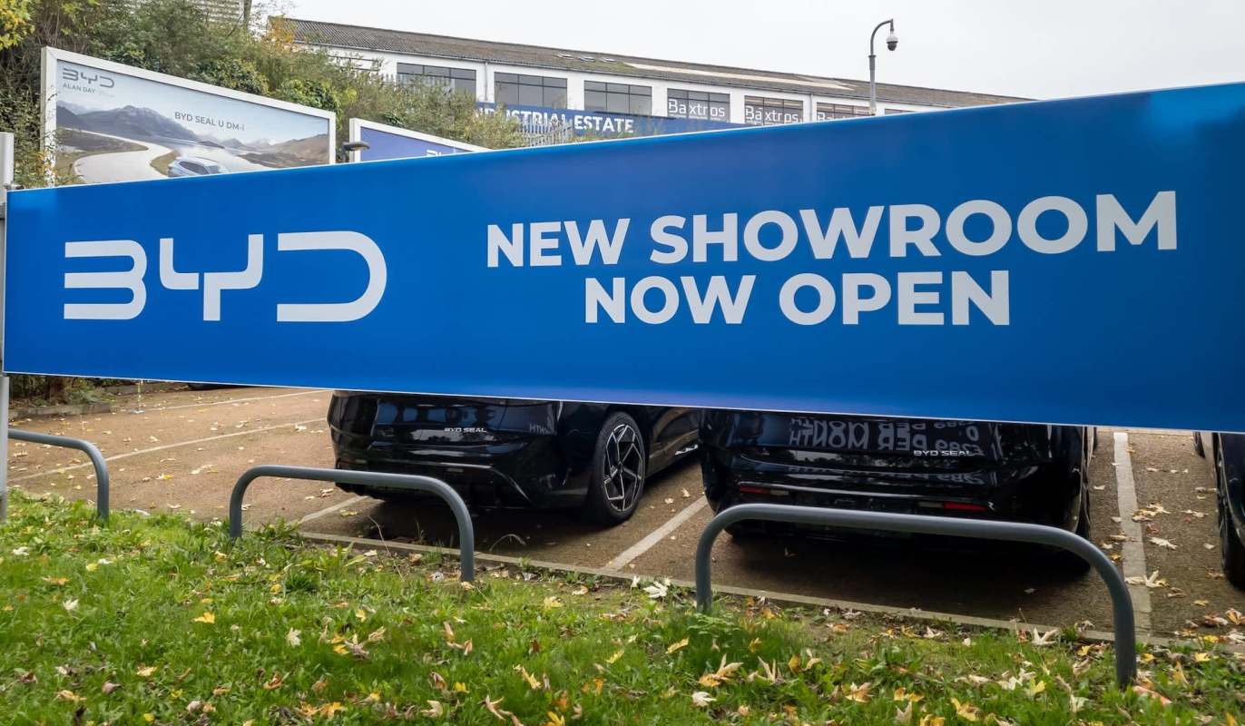 A large blue banner announces "NEW SHOWROOM NOW OPEN" at a BYD dealership, with parked cars visible below.