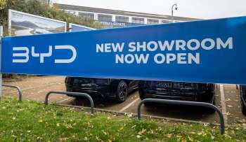 A large blue banner announces "NEW SHOWROOM NOW OPEN" at a BYD dealership, with parked cars visible below.