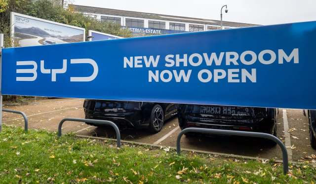 A large blue banner announces "NEW SHOWROOM NOW OPEN" at a BYD dealership, with parked cars visible below.