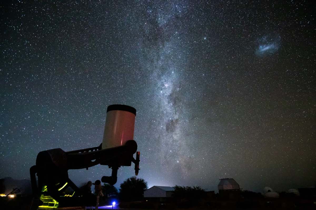 A telescope silhouetted against a starry sky, featuring the Milky Way and distant celestial objects.