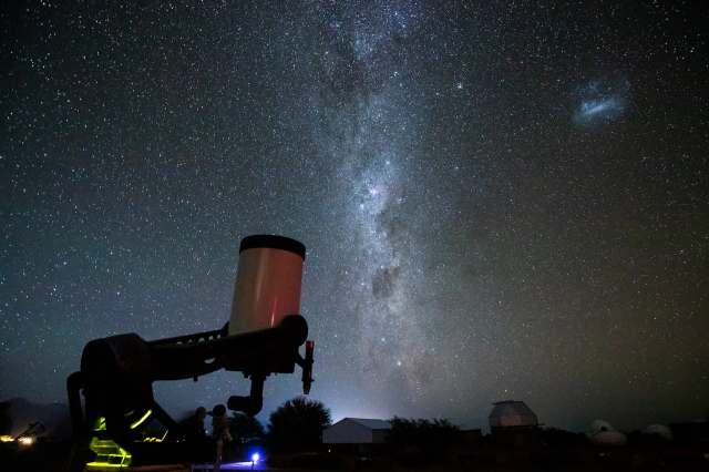 A telescope silhouetted against a starry sky, featuring the Milky Way and distant celestial objects.