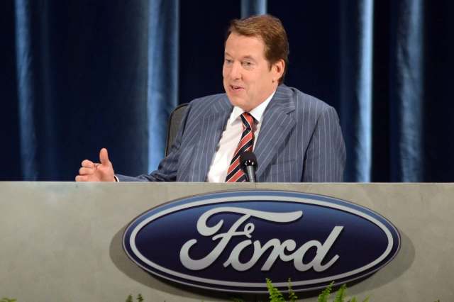 A man in a striped suit speaks at a podium with the Ford logo during a press event.