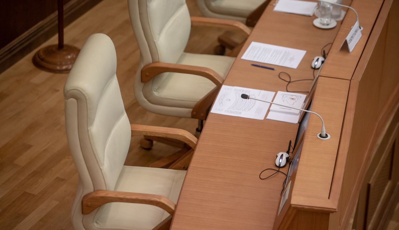 A close-up view of an empty committee chair beside documents and a microphone on a conference table.