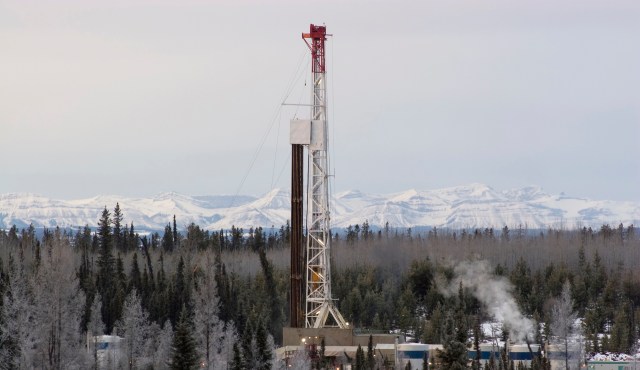 An oil drilling rig is positioned among trees with snow-capped mountains in the background.