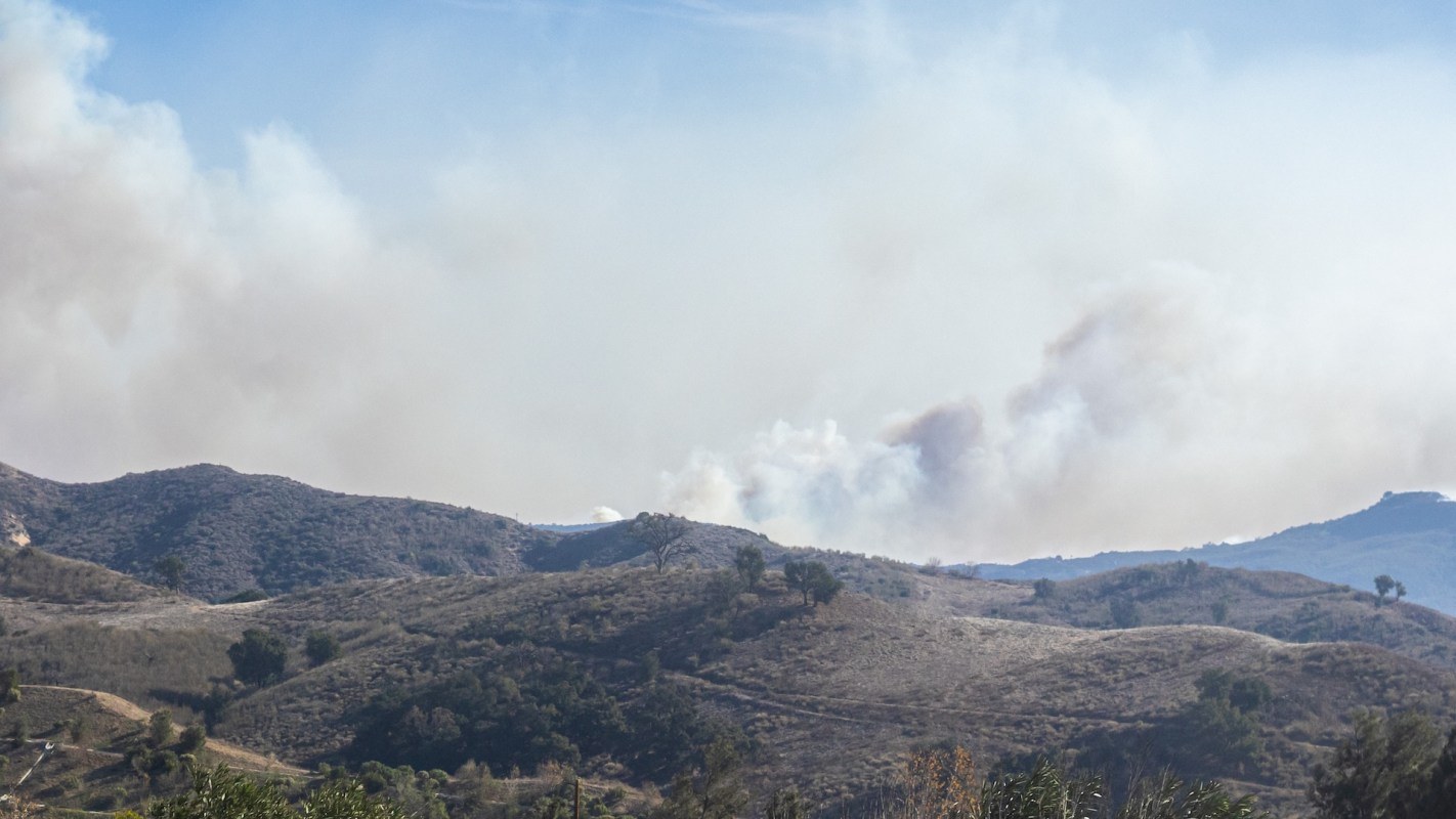 A hazy landscape showing rolling hills with smoke rising from the top, indicating a nearby wildfire.