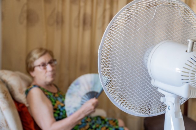 An elderly woman fanning herself as an electric fan blows toward her chair.