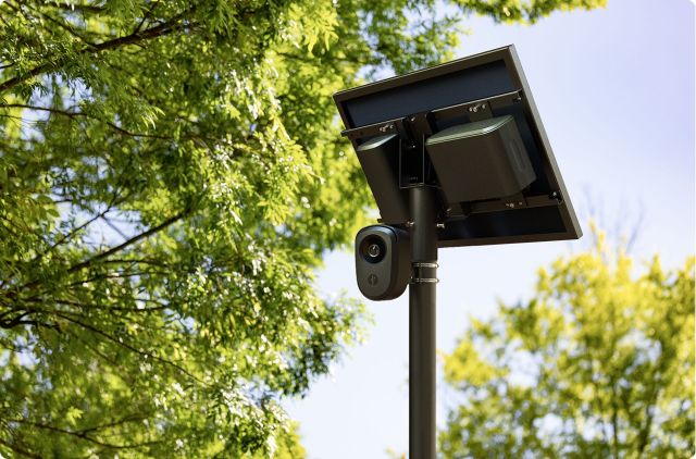 A solar-powered security camera mounted on a post, surrounded by green trees and a clear blue sky.