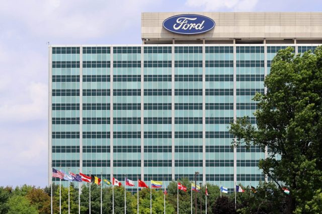 The Ford headquarters is seen behind multiple international flags and greenery under a partly cloudy sky.