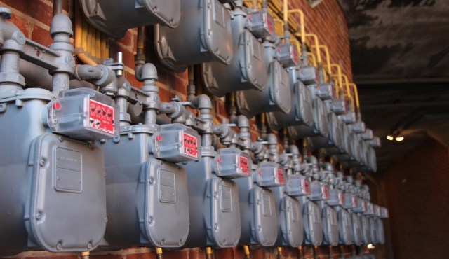 A row of gas meters attached to a brick wall, featuring grey casings and red dials.