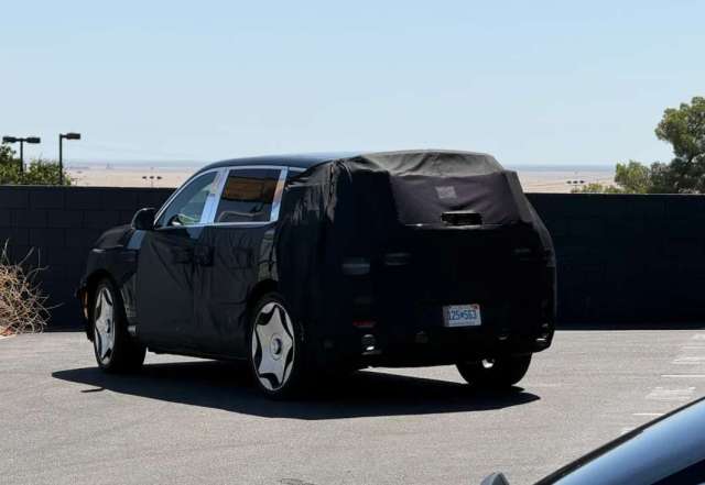 A blacked-out test vehicle is parked in a lot, partially obscured by covers and surrounded by sparse vegetation.