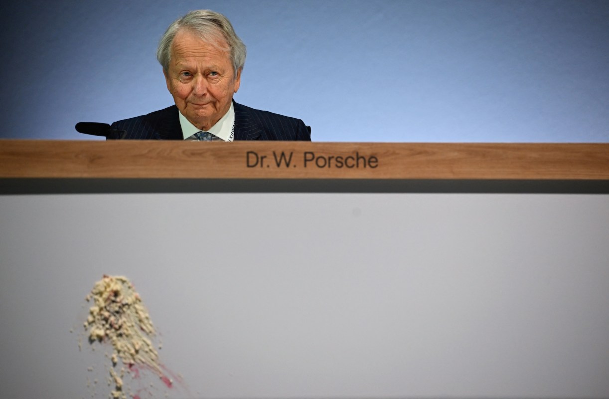 A man with gray hair in a suit sits behind a podium labeled "Dr. W. Porsche," with a cake stain on the side.