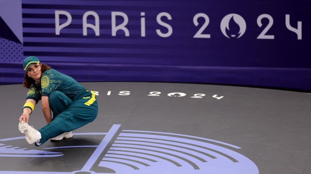 An athlete stretches on a competition mat with "Paris 2024" displayed in the background.