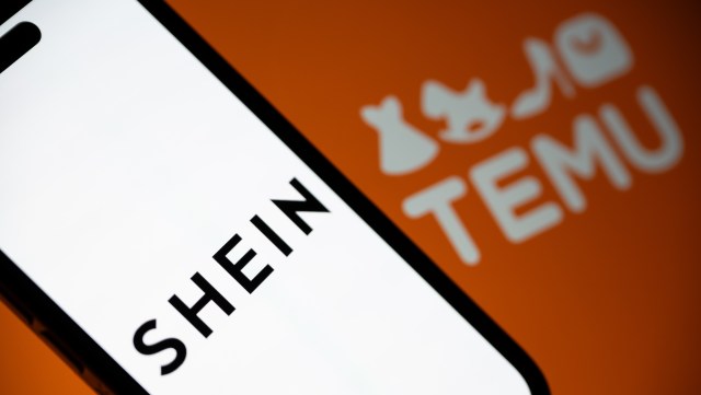 A close-up of a smartphone displaying the logo for SHEIN with the TEMU logo blurred in the background.