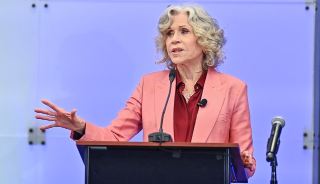 Jane Fonda, in a pink blazer, gestures while speaking at a podium with a microphone in a bright setting.