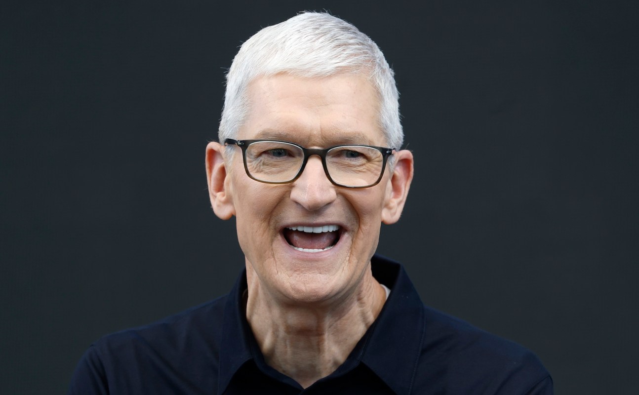 Apple CEO Tim Cook, with short gray hair and glasses, smiles while wearing a black shirt against a dark background.