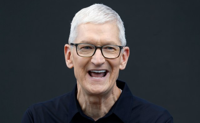 Apple CEO Tim Cook, with short gray hair and glasses, smiles while wearing a black shirt against a dark background.