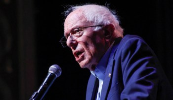 Bernie Sanders speaks passionately into a microphone on a dark stage.
